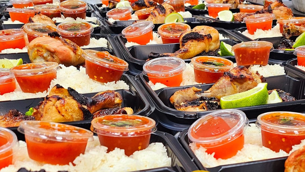 Dozens of Sticky Chikky catering meal boxes laid out side by side, each filled with charred chicken, sticky rice, a lime wedge, and a sealed cup of red sauce