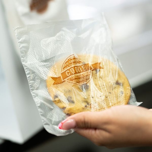 A hand holding a large chocolate chip cookie sealed in a clear bag with a branded COOKIES stamp