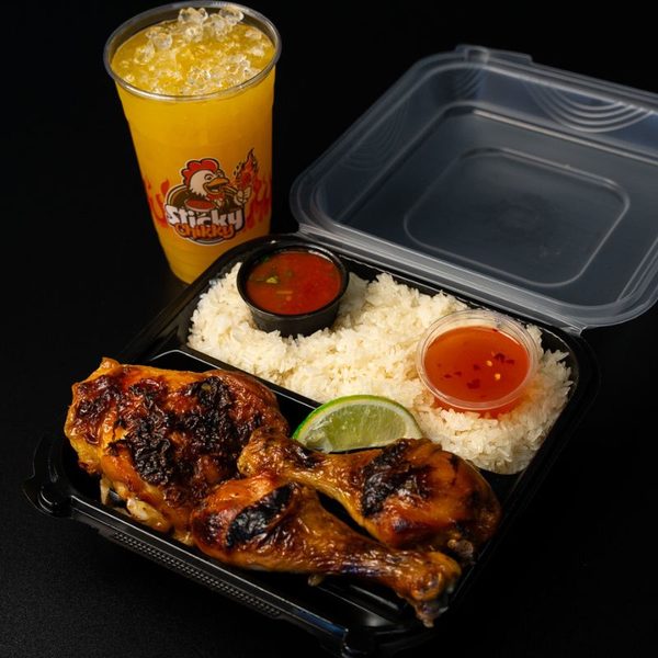 A full Sticky Chikky meal in a black takeout box: two charred drumsticks, sticky rice, two sauce cups, a lime wedge, and a branded orange drink with crushed ice alongside