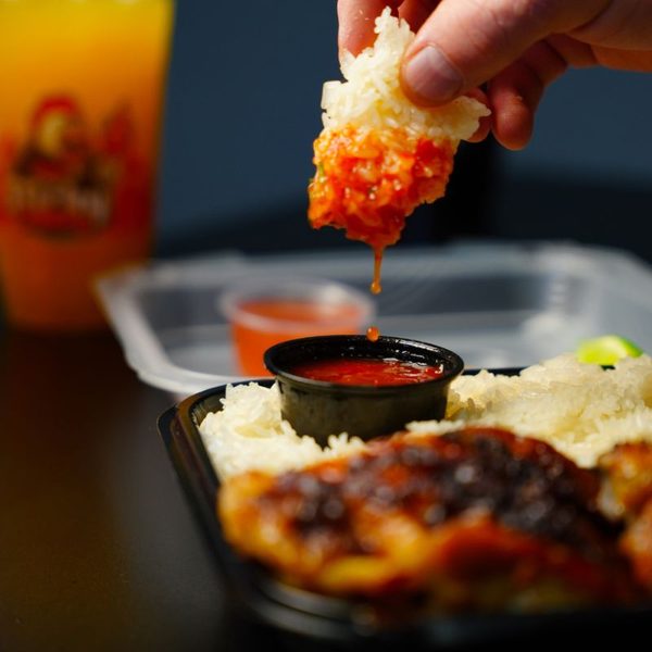 A hand dipping a clump of sticky rice coated in red sauce into a black sauce cup, with charred chicken and rice in a meal box below and a Sticky Chikky drink blurred in the background