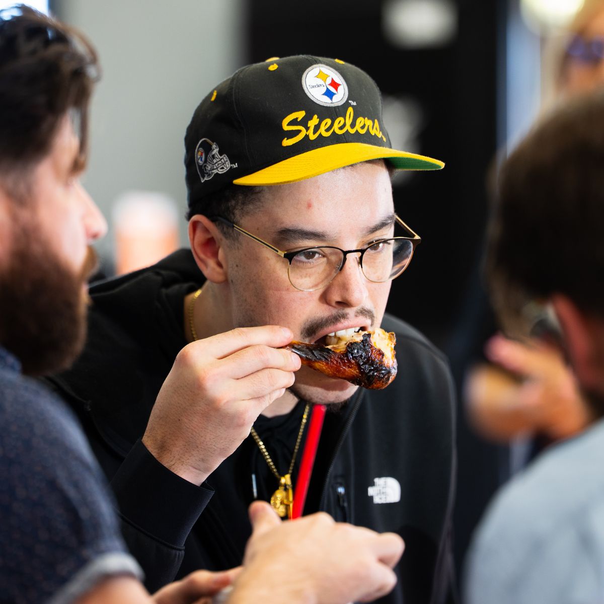 A man in a Steelers cap and glasses biting into a sticky glazed chicken drumstick at a Sticky Chikky tasting event
