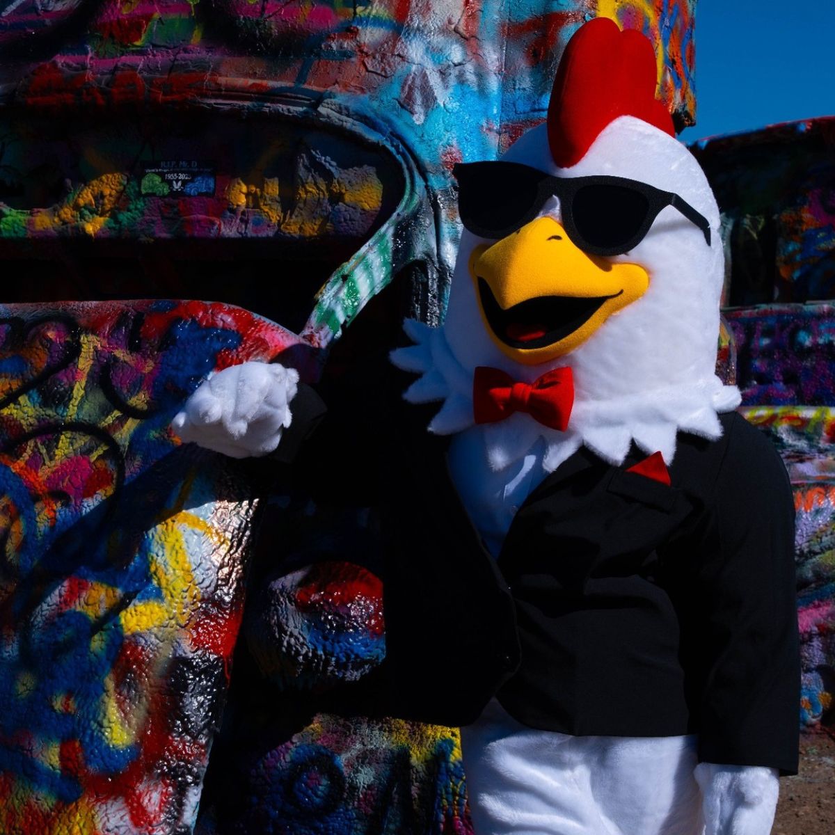 The Sticky Chikky chicken mascot in sunglasses leaning against a graffiti-covered car at Cadillac Ranch, Amarillo