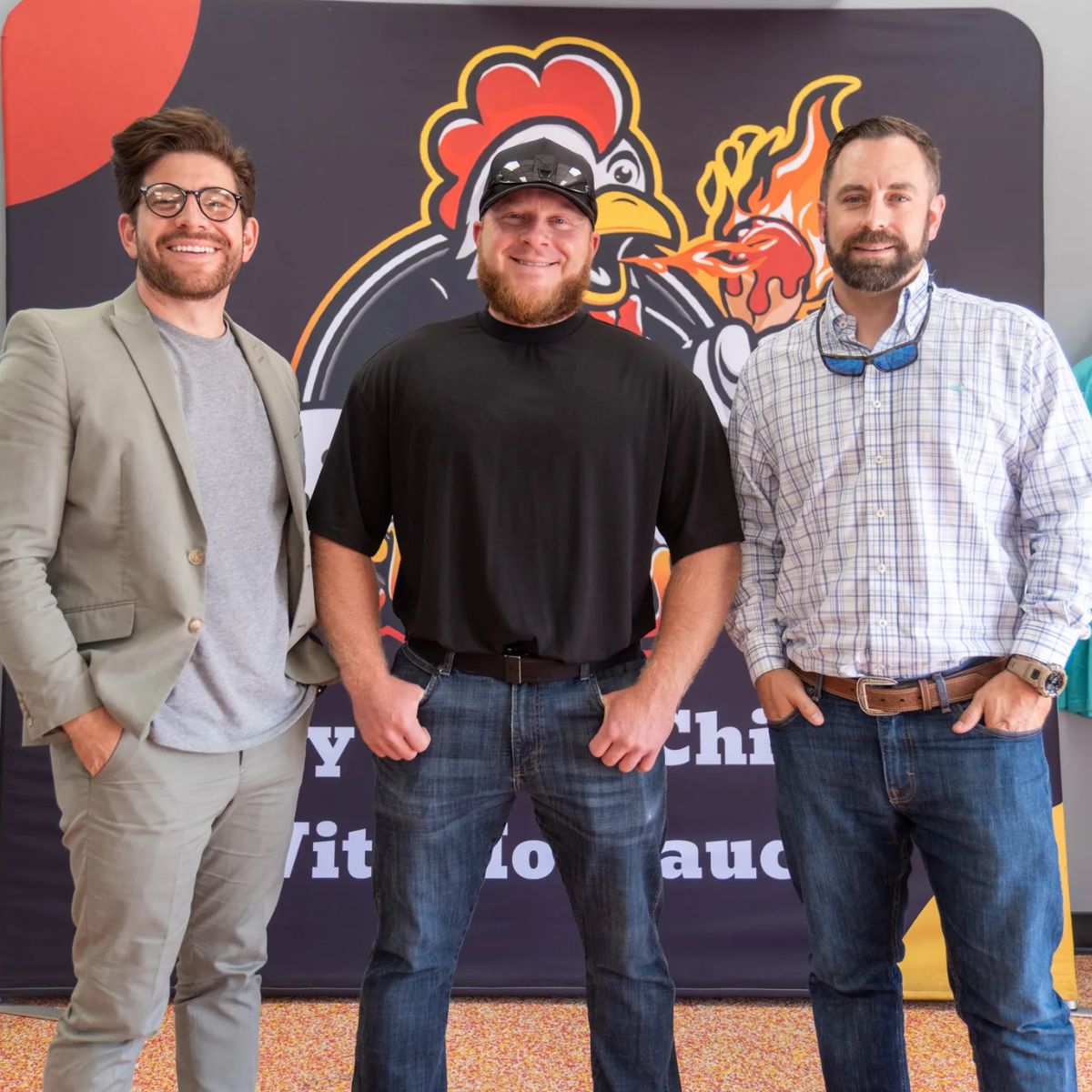 Three men smiling together in front of a Sticky Chikky branded banner at a restaurant event
