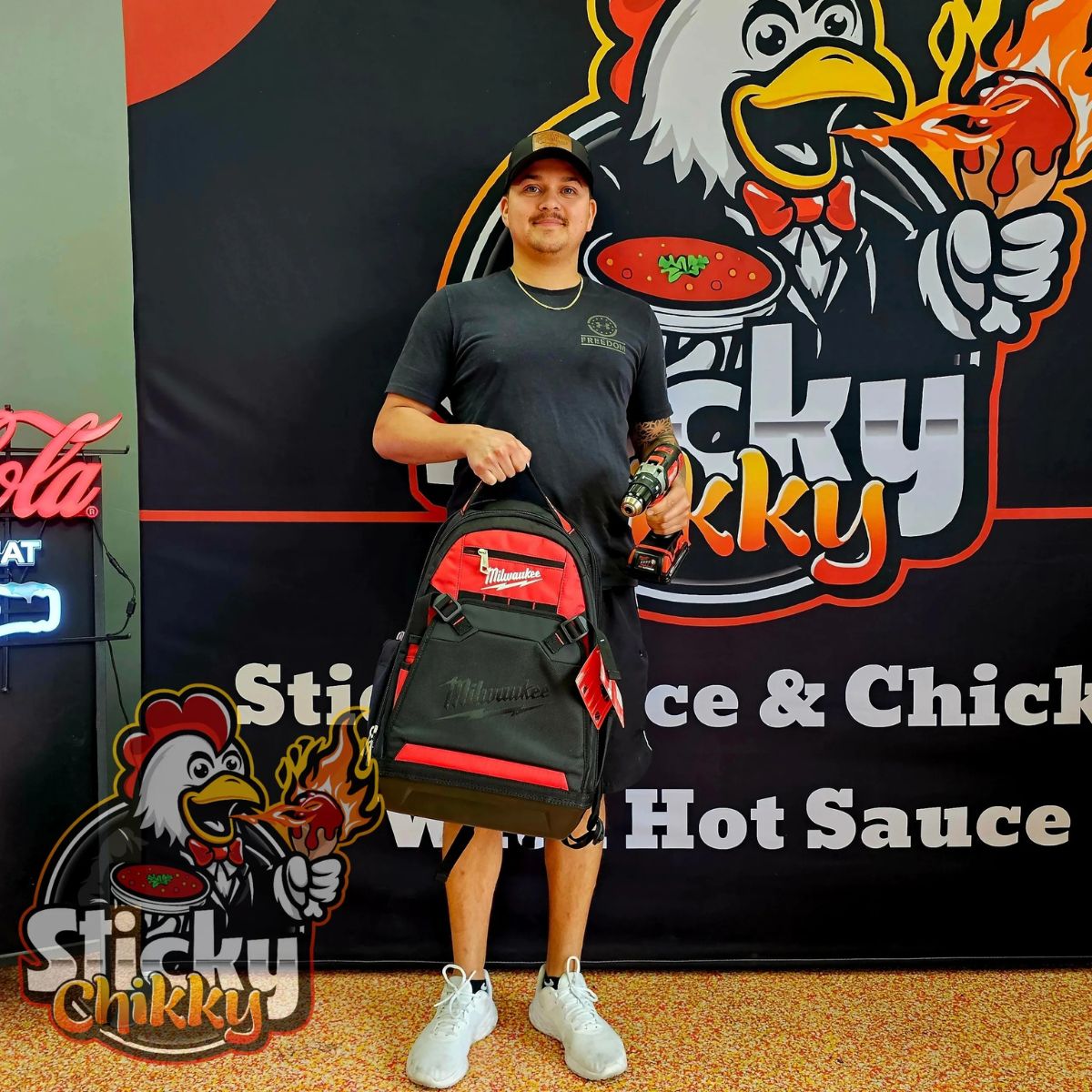 A man holding a Milwaukee tool bag and drill in front of a Sticky Chikky branded banner — a giveaway winner at the restaurant