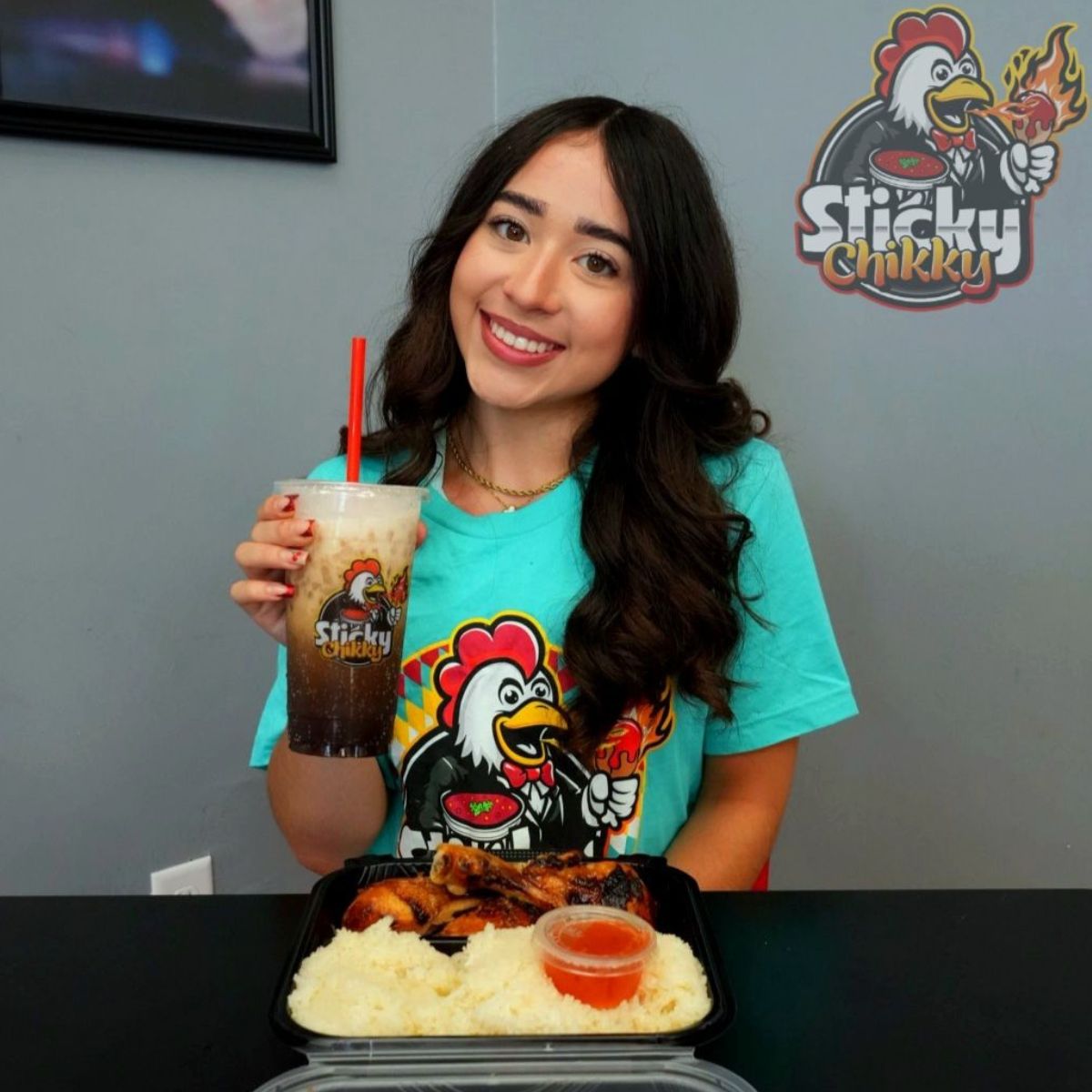 A smiling woman in a teal Sticky Chikky t-shirt holding an iced drink with a chicken and rice meal tray in front of her inside the restaurant