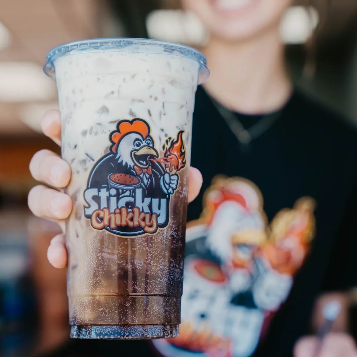 A hand holding a large Sticky Chikky branded cup filled with a creamy iced drink, with the restaurant mascot logo on the cup