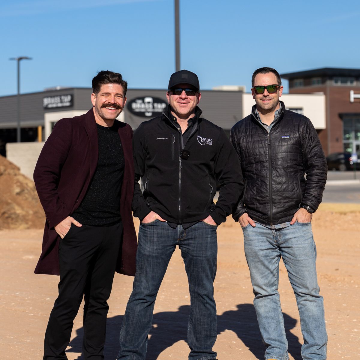 Three men smiling together on an empty lot at a future Sticky Chikky construction site, with retail buildings visible in the background