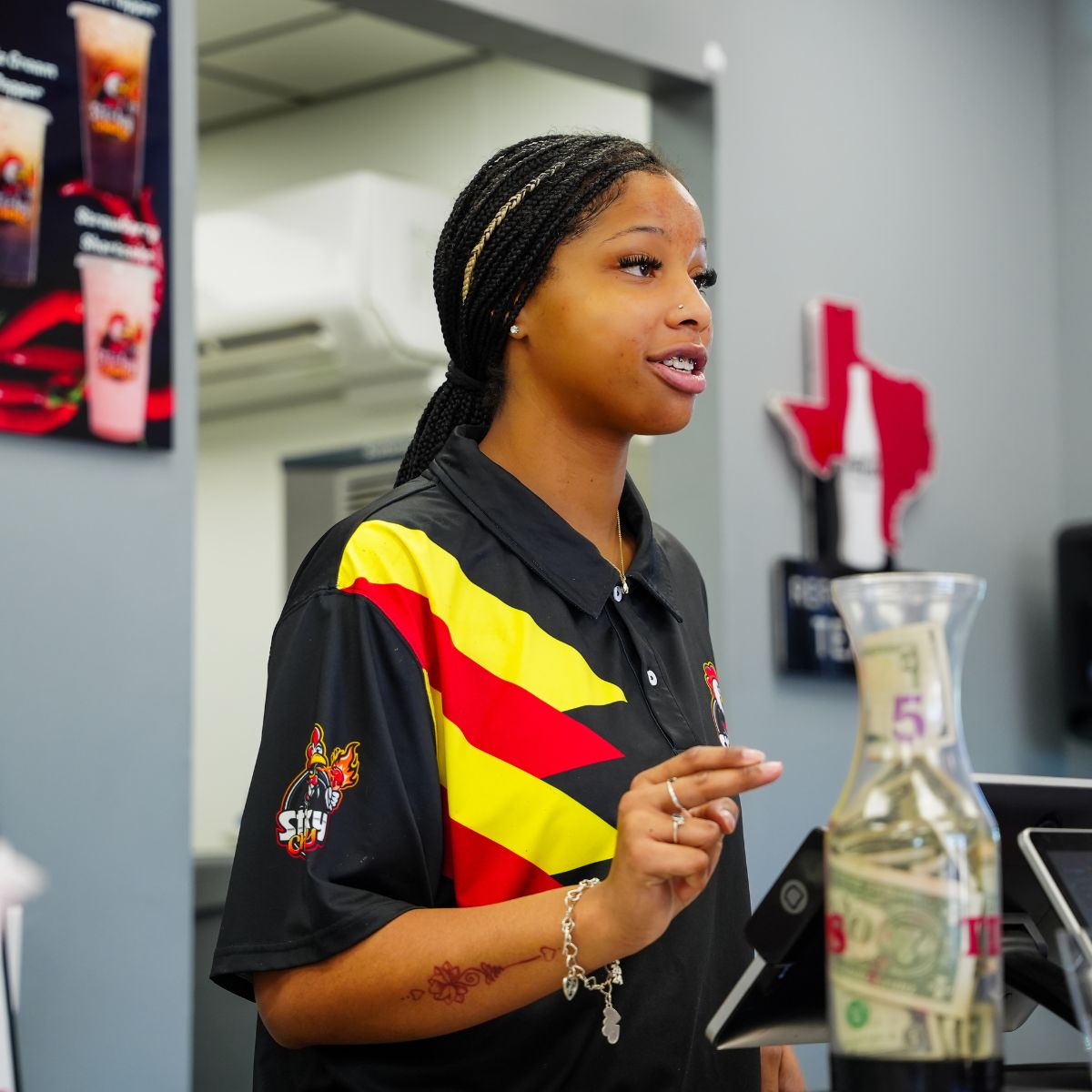 A Sticky Chikky team member in the black-and-yellow branded uniform at the register, with a tip jar and Texas-shaped décor on the counter behind her