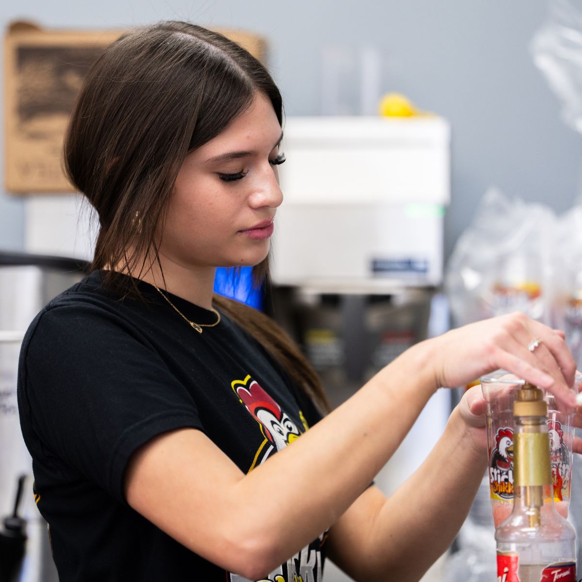 A Sticky Chikky team member in a black branded t-shirt preparing a drink behind the counter, pouring syrup into a cup with the Sticky Chikky logo