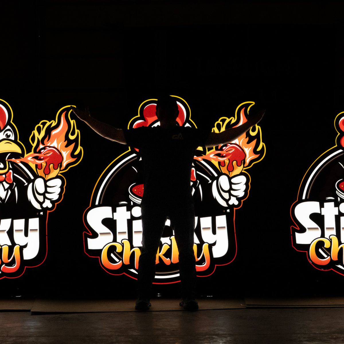 A silhouetted figure standing arms outstretched in front of three illuminated Sticky Chikky logo light-box signs glowing against a dark background