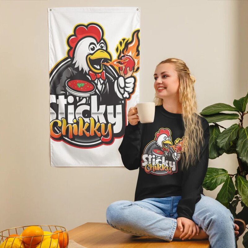 Woman sitting cross-legged wearing a black Sticky Chikky crew neck sweatshirt with the brand logo on the chest, large brand banner visible behind her