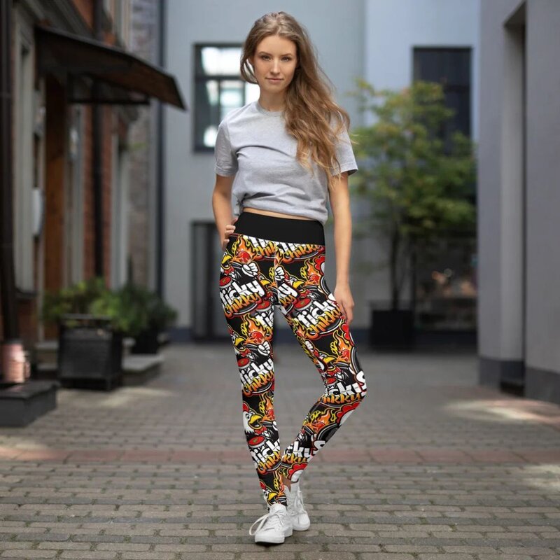Woman standing on a cobblestone street wearing all-over print yoga leggings with a repeated Sticky Chikky logo and flame pattern in red, orange, yellow, and black, with a black waistband